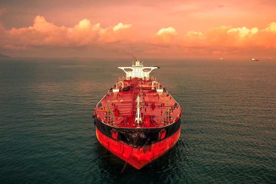 A large red dry bulk cargo ship in calm open waters under a dramatic orange sky at sunset, heading straight forward