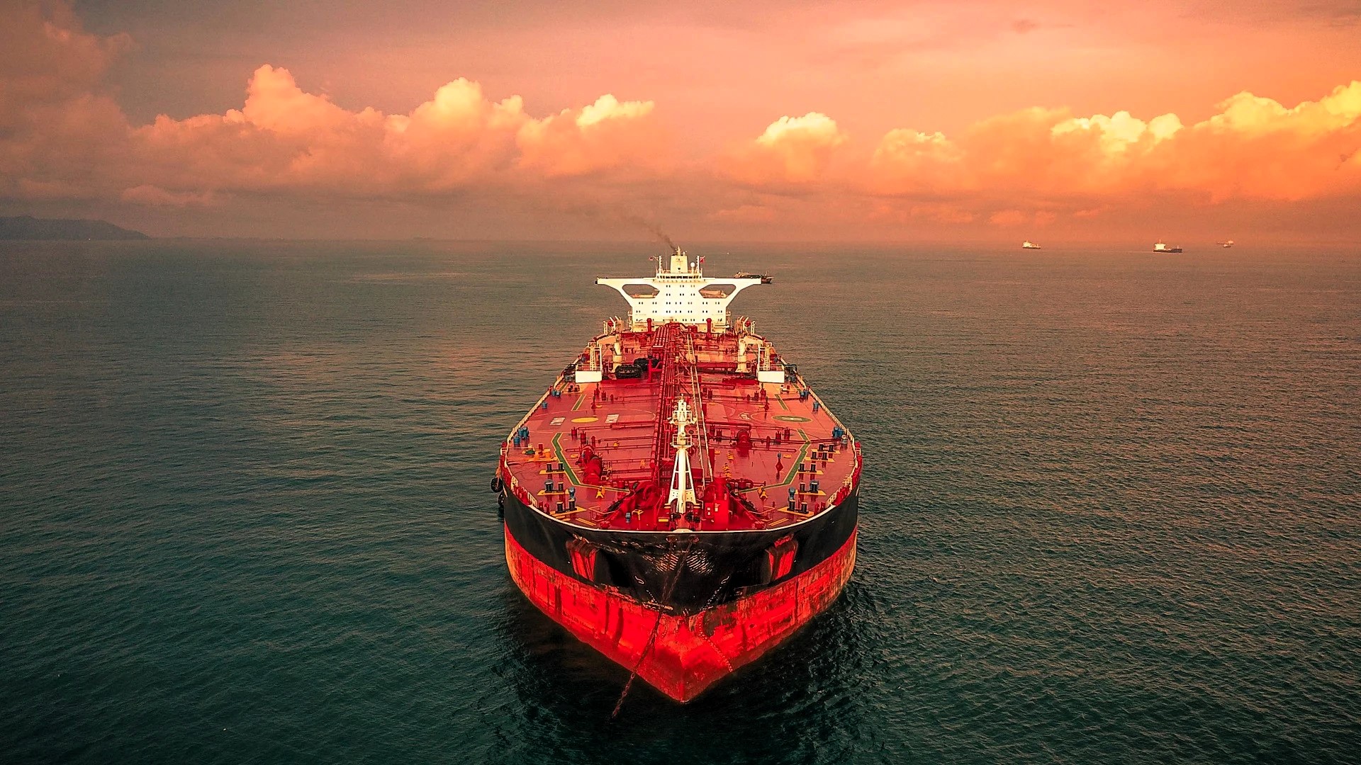 A large red dry bulk cargo ship in calm open waters under a dramatic orange sky at sunset, heading straight forward