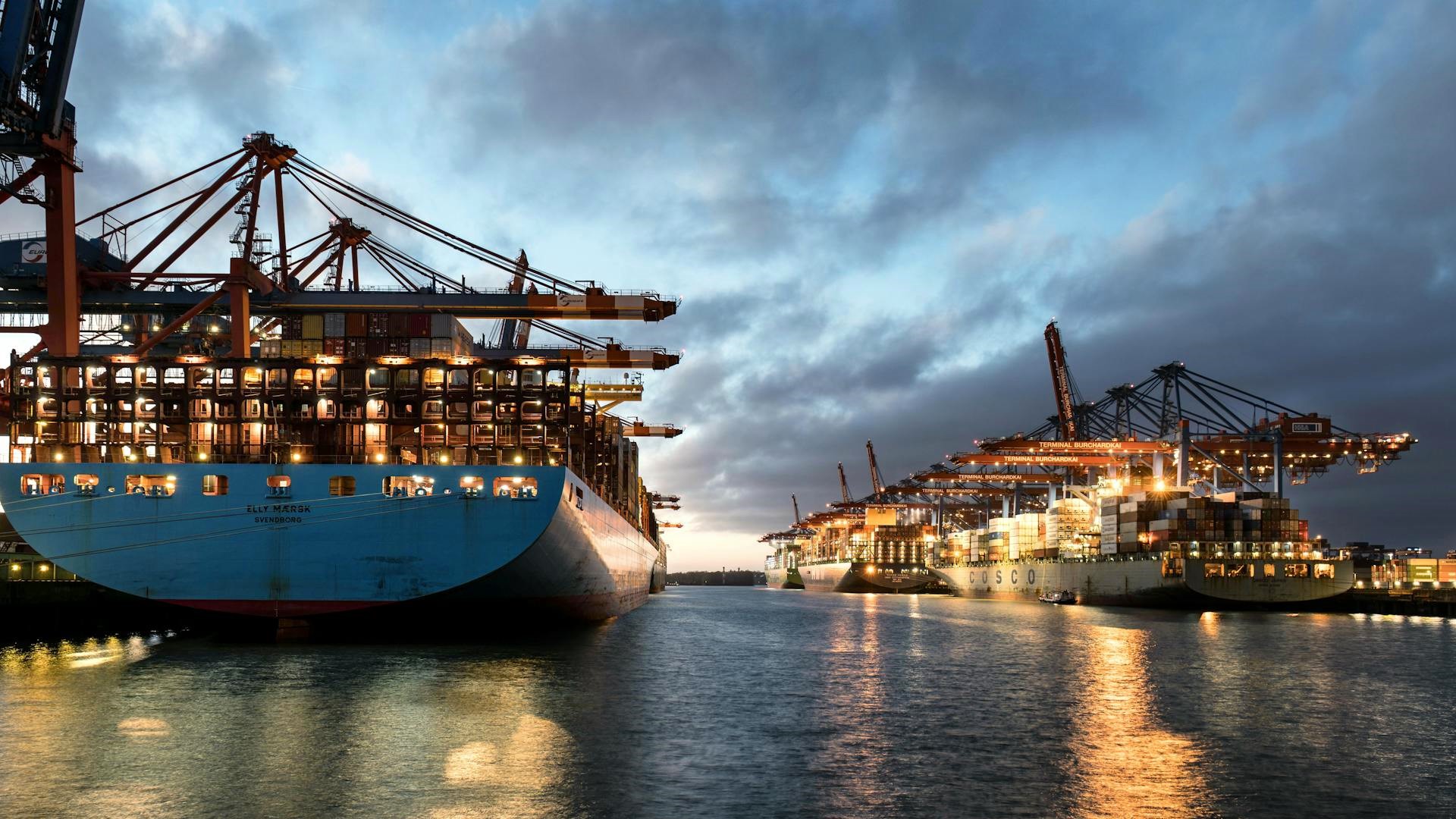 global shipping disruptions_geo-trends.eu_A bustling shipping terminal at dusk, featuring large cargo ships loaded with containers, illuminated by bright industrial lights, with cranes in the background under a cloudy sky