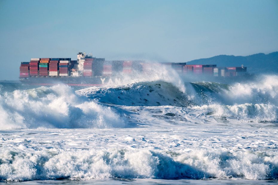 A cargo ship sailing in the background of a turbulent ocean with large waves crashing in the foreground, under clear blue skies. The scene highlights the challenges faced by the shipping industry due to extreme weather conditions caused by climate change