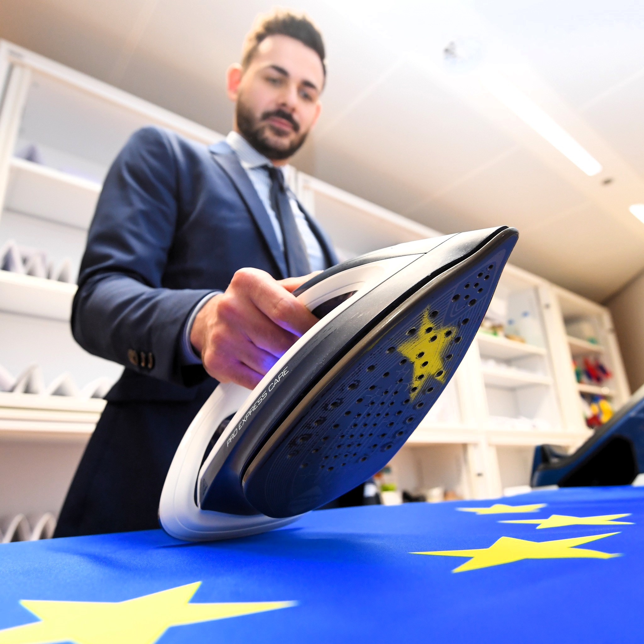 A man in a suit holds an iron with a yellow star imprinted on its soleplate, pressing it onto an EU flag. The image symbolizes efforts to “fix” or “smooth out” Europe’s challenges, reflecting ongoing economic and political turmoil