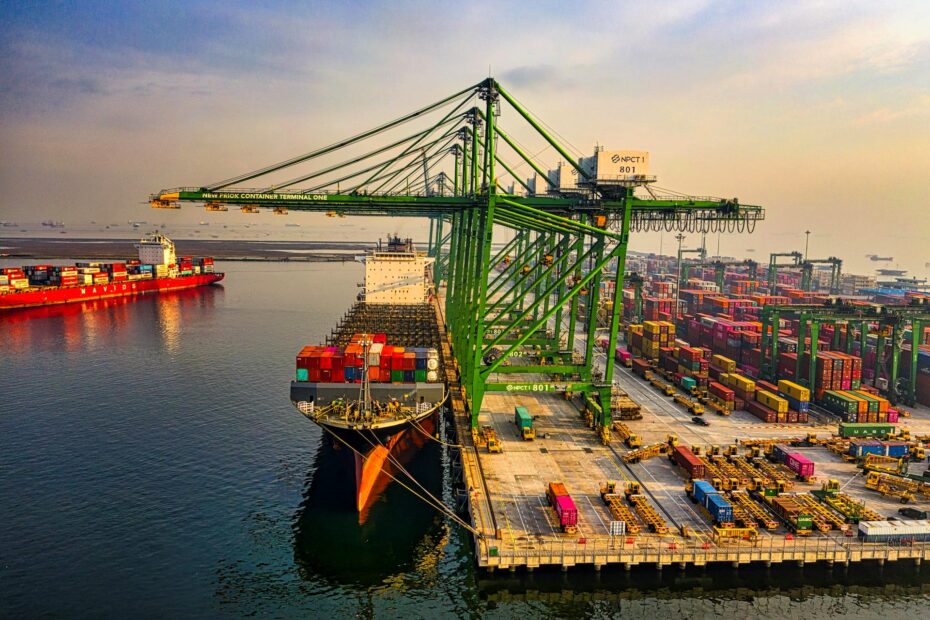 dry bulk market_Aerial view of a busy container terminal in North Jakarta, Indonesia, captured by Tom Fisk. A large container ship is docked at the terminal, with towering green gantry cranes unloading and loading containers. The port is filled with neatly stacked multicolored containers, trucks, and equipment actively operating. The calm sea reflects the golden hues of the setting sun, while multiple cargo ships are visible in the distance