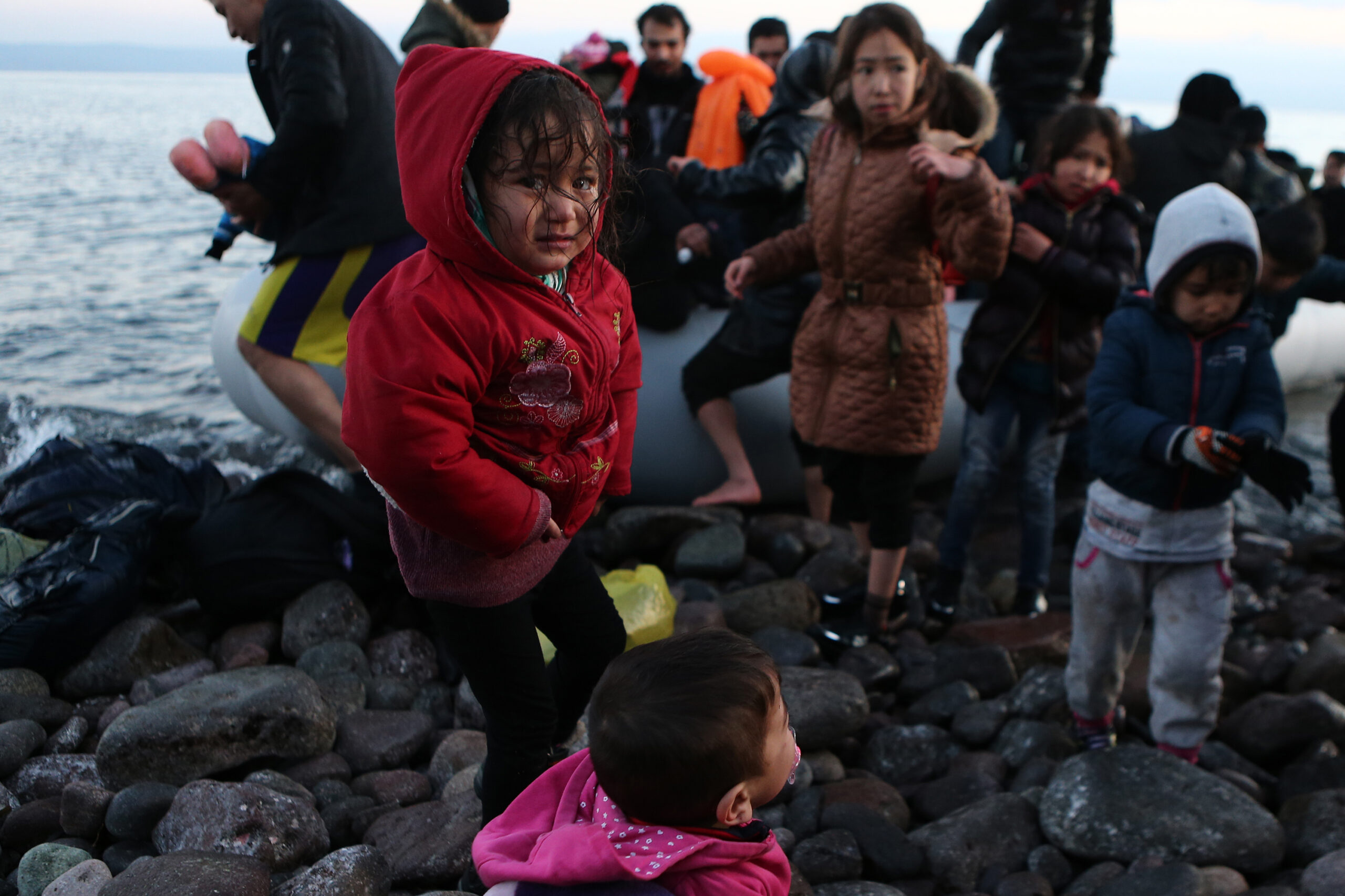 A group of migrants, including young children, arrive on a rocky shore after crossing the sea in an inflatable boat. A little girl in a red jacket stands in the foreground, her face wet and filled with emotion. Behind her, other children and adults, dressed in warm but worn clothing, step onto the shore, some looking exhausted, others scanning their surroundings. The cold, dark water and scattered belongings emphasize the perilous nature of their journey