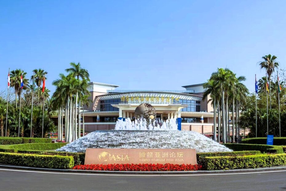 The Boao Forum for Asia conference center with a large fountain in front, a golden globe sculpture, palm trees, and multiple international flags under a clear blue sky