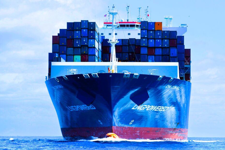 A large CMA CGM container ship, LNG-powered, navigating through open waters with a full load of multicolored shipping containers stacked high on its deck. The ship’s bow cuts through the ocean, with American and Chinese flags visible atop the bridge
