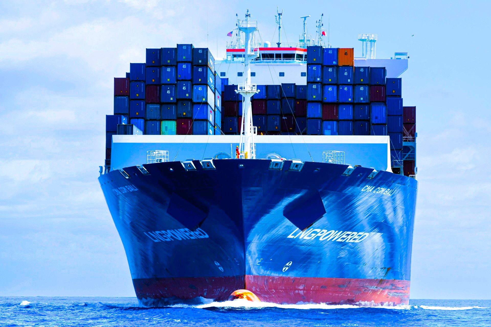A large CMA CGM container ship, LNG-powered, navigating through open waters with a full load of multicolored shipping containers stacked high on its deck. The ship’s bow cuts through the ocean, with American and Chinese flags visible atop the bridge