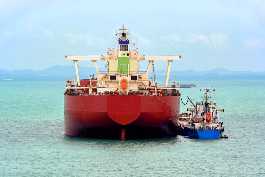 A large cargo vessel receiving fuel from a bunker barge at sea. The fueling process, known as bunkering, is susceptible to scams like the Cappuccino Effect, where compressed air is injected into fuel to inflate its volume fraudulently