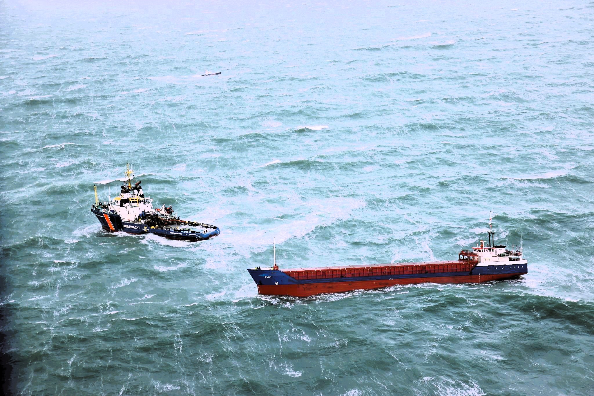 Aerial view of a cargo ship and a coast guard vessel in rough seas. The cargo ship, painted red and blue, moves through choppy waters, while the coast guard ship, marked “KUSTWACHT,” appears to be escorting or monitoring it. The vast ocean in the background emphasizes the maritime industry’s vulnerability to geopolitical instability