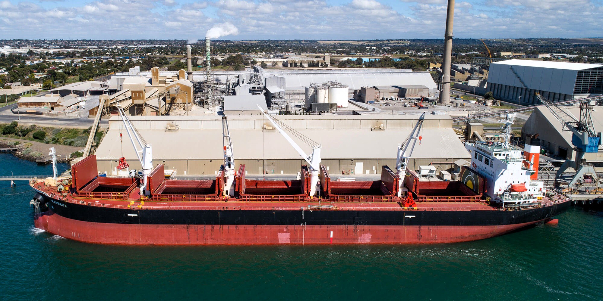 A large dry bulk cargo ship docked at an industrial port, with open cargo holds and cranes positioned for loading or unloading. The surrounding infrastructure includes warehouses, chimneys, and conveyor systems, indicating an active shipping hub