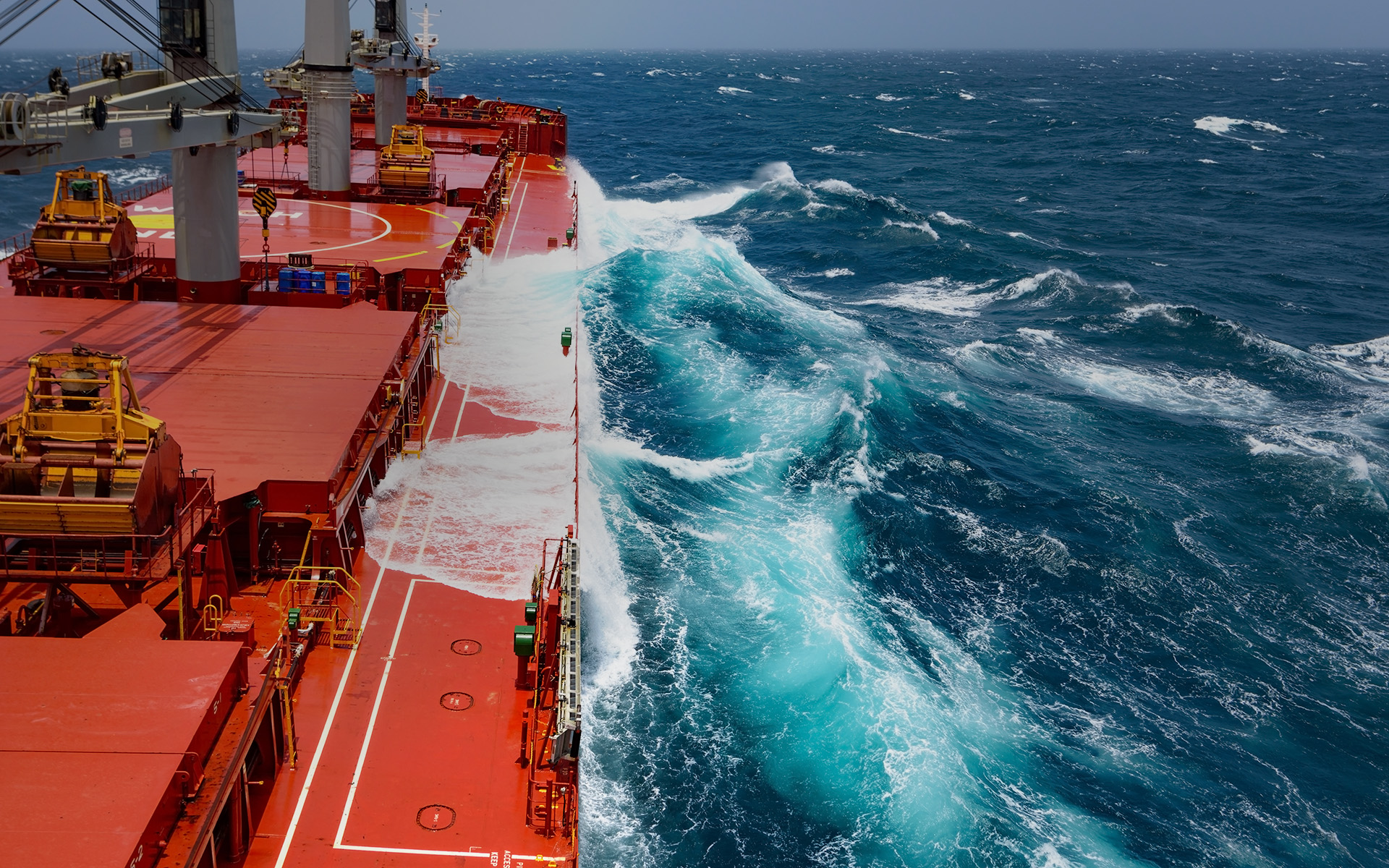 A large red bulk carrier ship navigating through rough ocean waters, with waves crashing against its hull. The vessel’s deck features cranes and equipment used for handling dry bulk cargo such as coal or iron ore. The deep blue sea contrasts with the ship's red structure, capturing the challenges of maritime trade