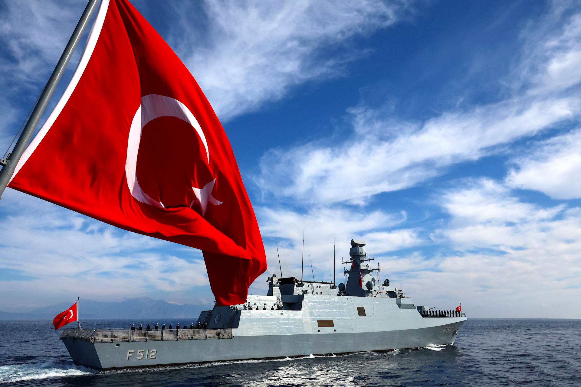 A Turkish naval vessel with the designation “F-512” sails on calm waters under a bright blue sky with scattered clouds. A large Turkish flag waves in the foreground, while another flag is visible at the ship’s stern. Navy personnel stand in formation along the deck. Mountains are faintly visible in the background