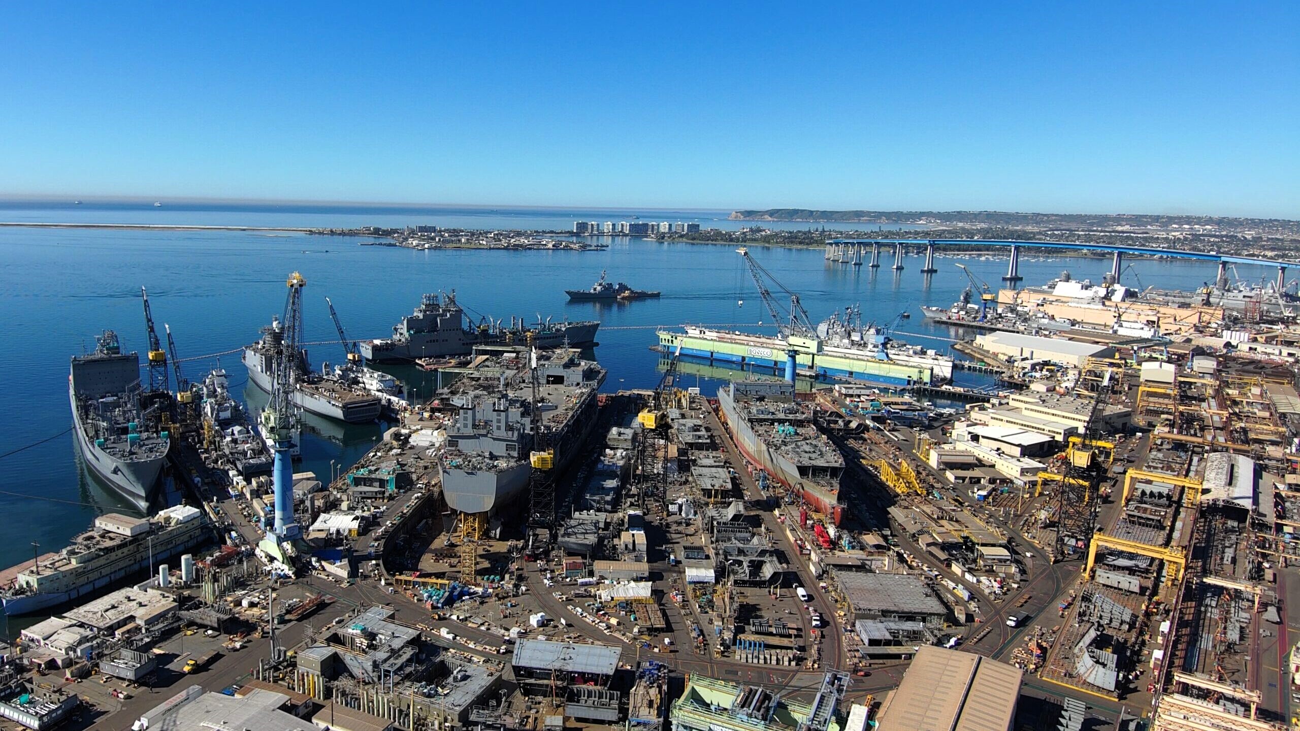 General Dynamics NASSCO shipyard with multiple vessels under construction and repair, featuring cranes, industrial equipment, and extensive shipping infrastructure. The shipyard is located by a harbor with ships and a bridge in the background