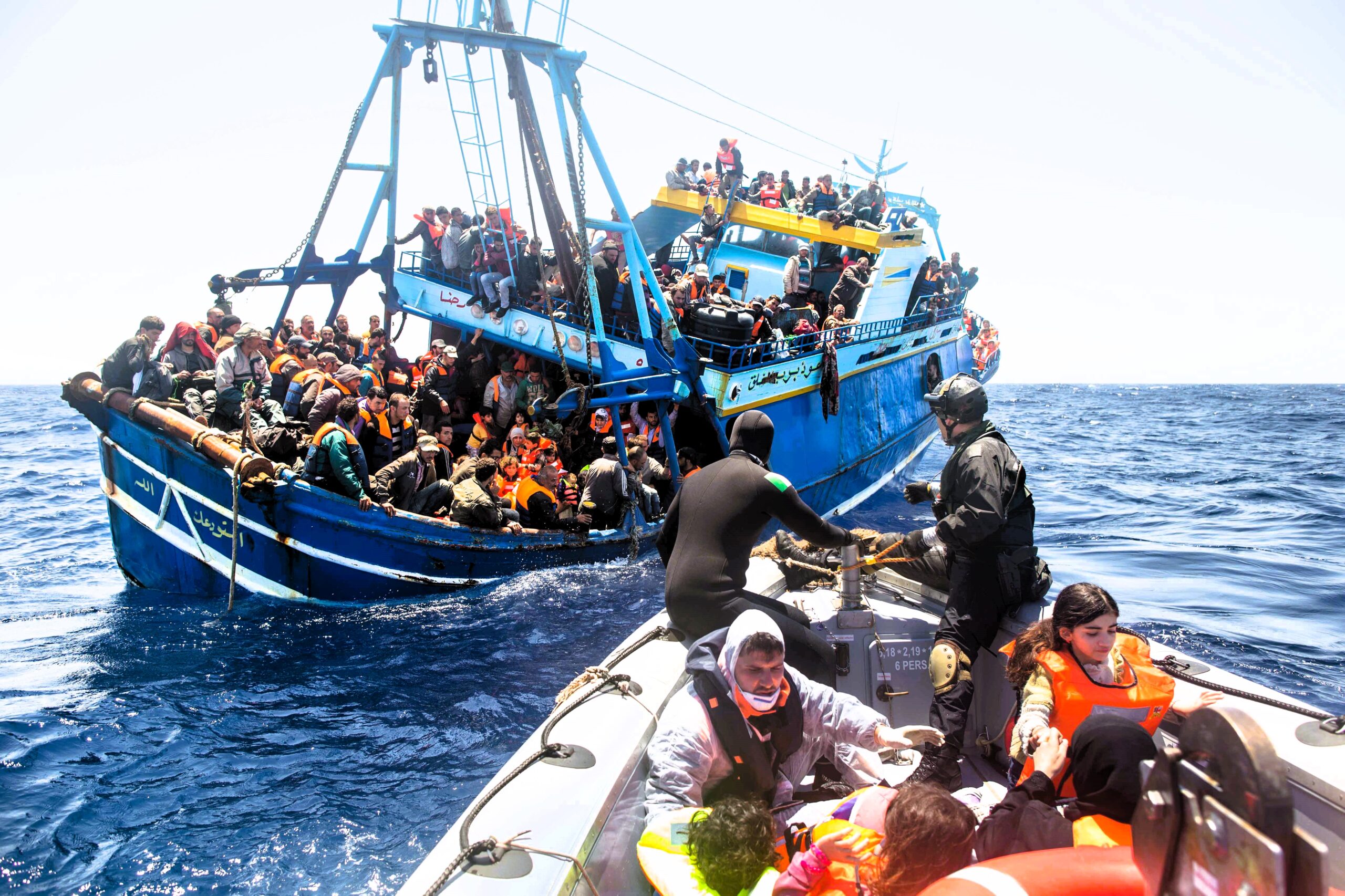 A heavily overcrowded blue fishing boat carrying migrants in distress on the open sea. Many passengers wear life jackets, while others cling to the vessel. A section of the boat appears damaged. Nearby, members of the Italian Coast Guard, dressed in tactical and rescue gear, assist people onto a smaller rescue boat. The surrounding sea is calm under a bright sky, highlighting the urgency of the rescue operation
