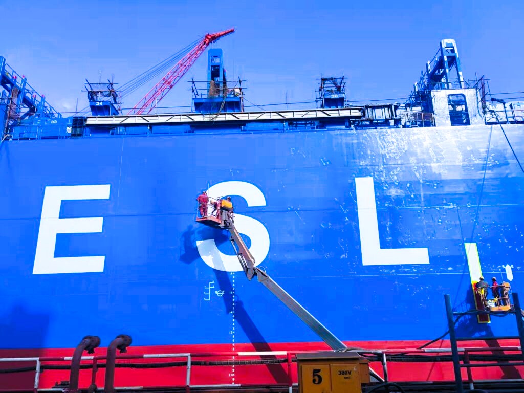 Workers repaint the hull of a large Emirates Shipping Line vessel at a shipyard, showing giant white letters “ESL” on a blue and red background, under a clear blue sky