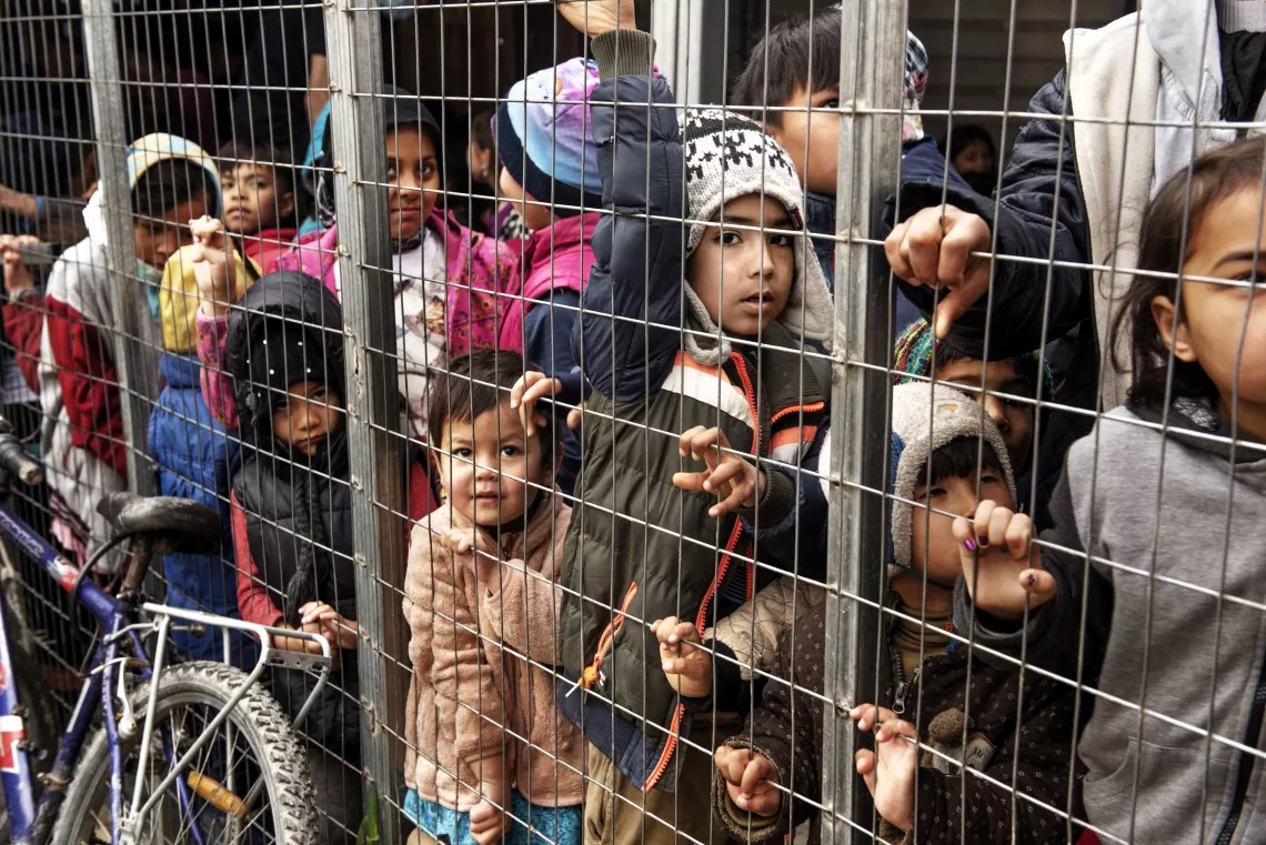 A group of refugee and migrant children stand behind a metal fence inside the Reception and Identification Centre in Moria, Lesvos, Greece, wearing winter clothes and looking out with somber expressions. A bicycle is seen in the foreground