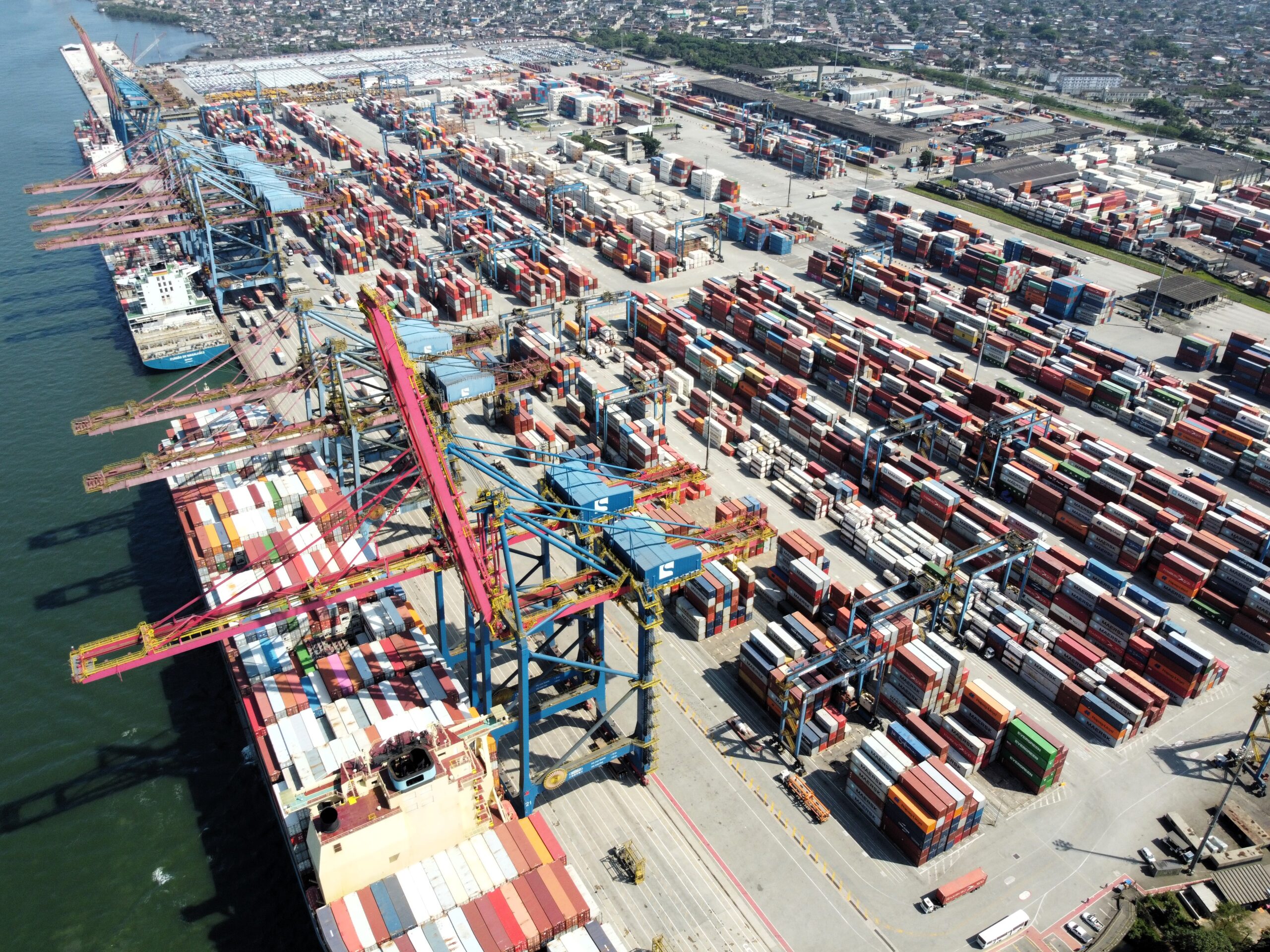 Aerial view of the Port of Santos in Brazil showing container ships docked alongside multiple gantry cranes and densely stacked multicolored shipping containers across a vast terminal, with adjacent urban infrastructure in the background