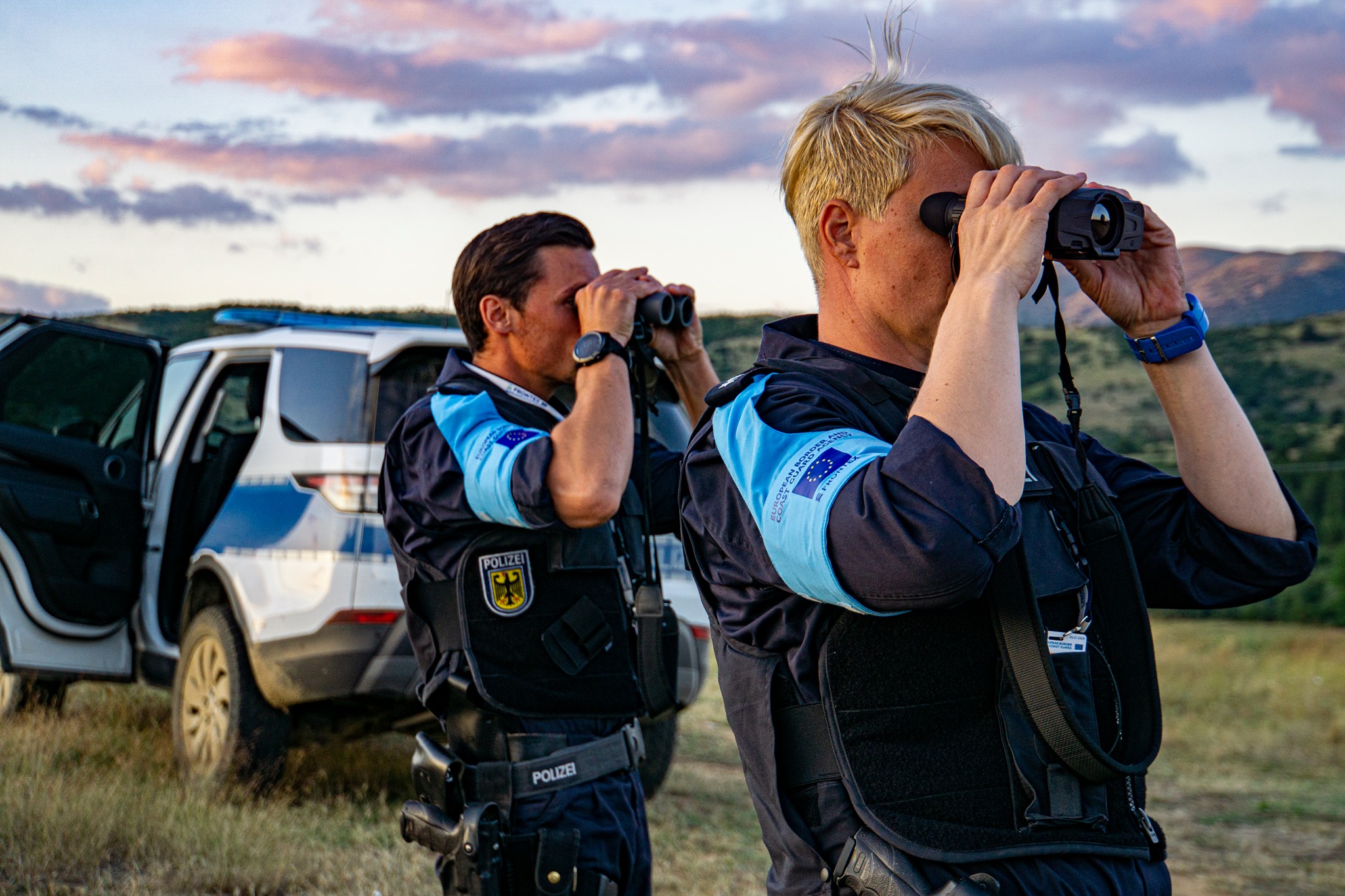 Two European border officers, wearing uniforms marked with both EU and national police insignia, are standing in front of a patrol vehicle with the doors open, using binoculars to survey the landscape during sunset. The terrain is open and rural, with mountains in the background and a partly cloudy sky. The scene captures a moment of coordinated field monitoring as part of EU border management operations