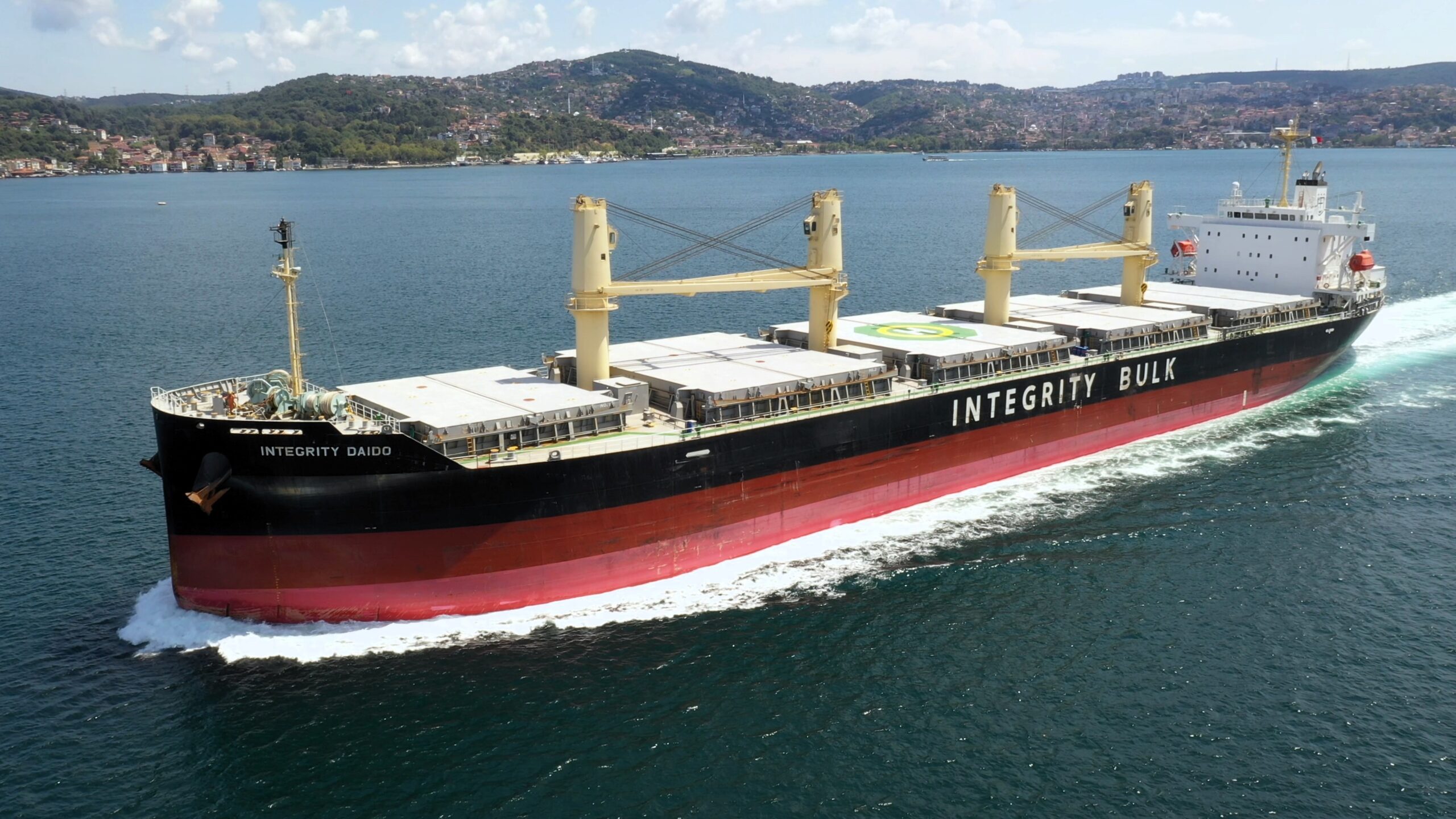 A bulk carrier vessel named “INTEGRITY DAIDO” sailing at sea, with cranes and cargo holds visible, operated by Integrity Bulk, passing near a coastal town with hills in the background