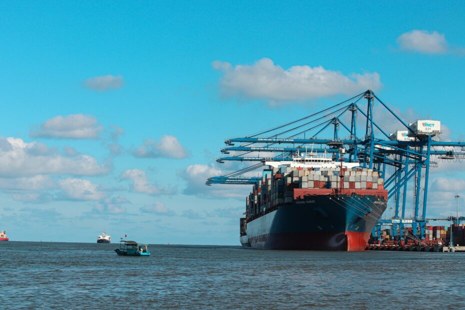 Large container ship docked at a busy industrial port with cranes loading cargo under a bright blue sky, representing maritime transport and logistics infrastructure