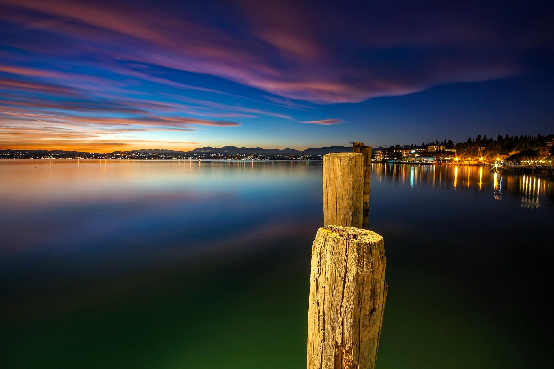 Sunset view of the port in Sirmione, Italy, with wooden posts in the foreground and lights along the shoreline
