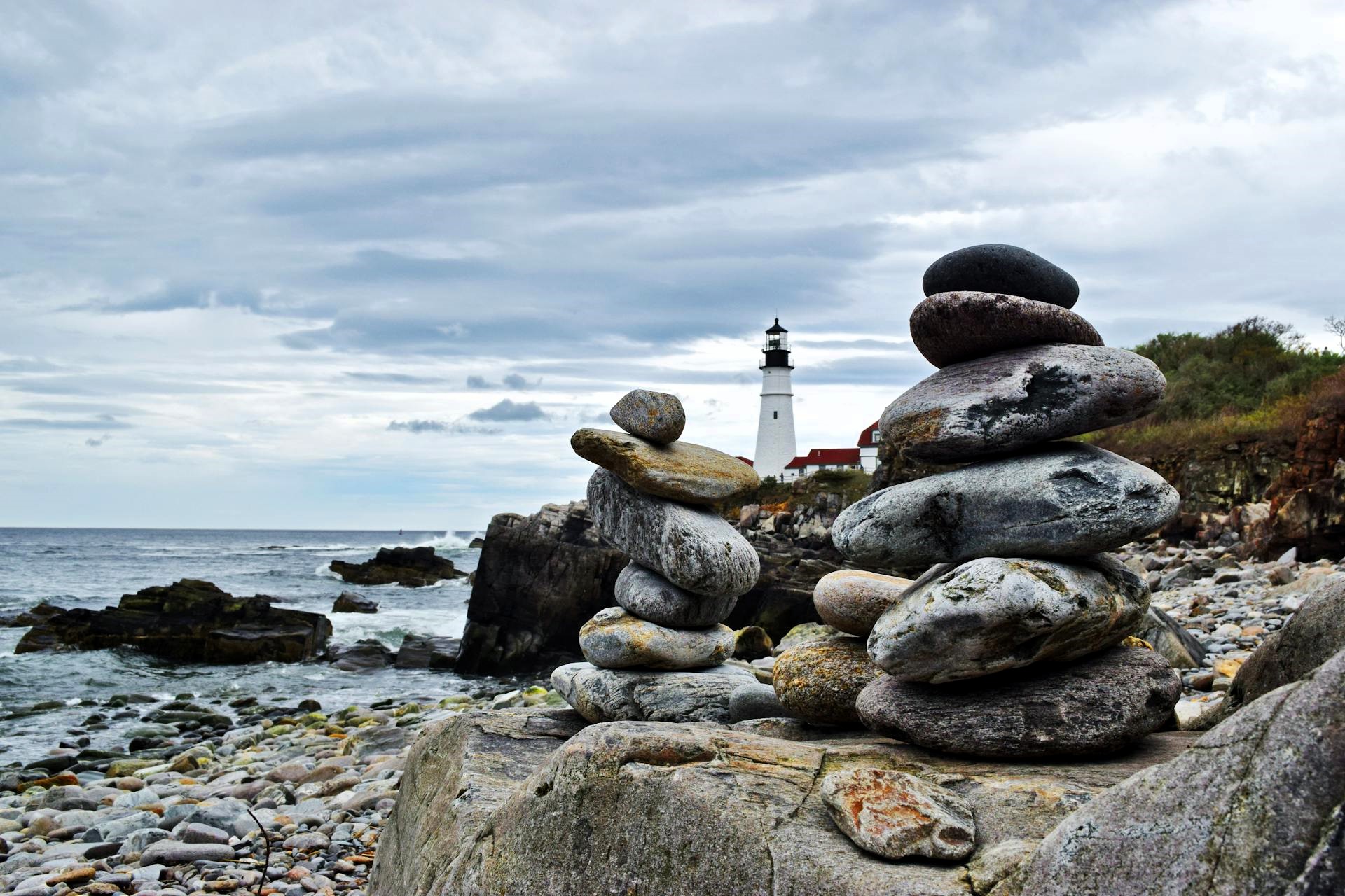 Stacked stones on a rocky shore with lighthouse in background, symbolizing fragile balance amid turbulent maritime markets