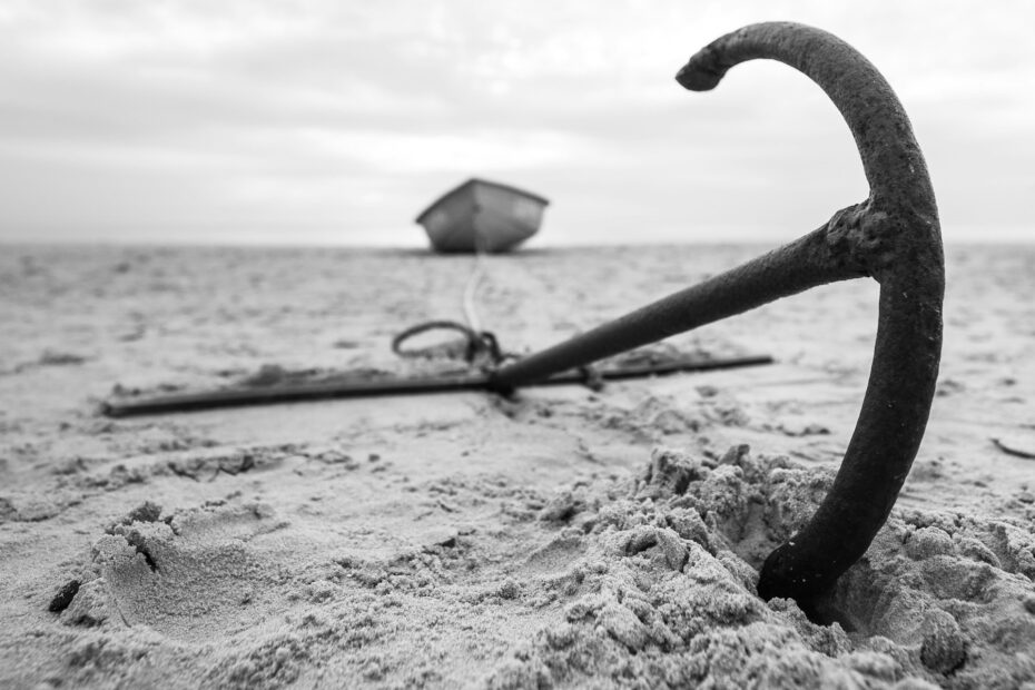 Rusty anchor in sand with blurred boat in background, symbolizing instability and disruption in global maritime trade