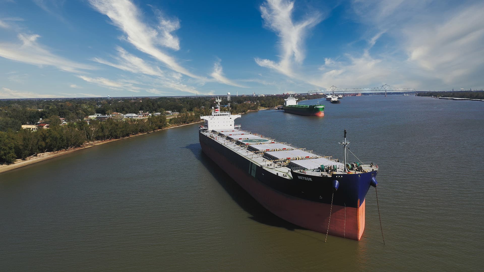 A large dry bulk carrier anchored near the shore, with other cargo ships visible along a wide river channel