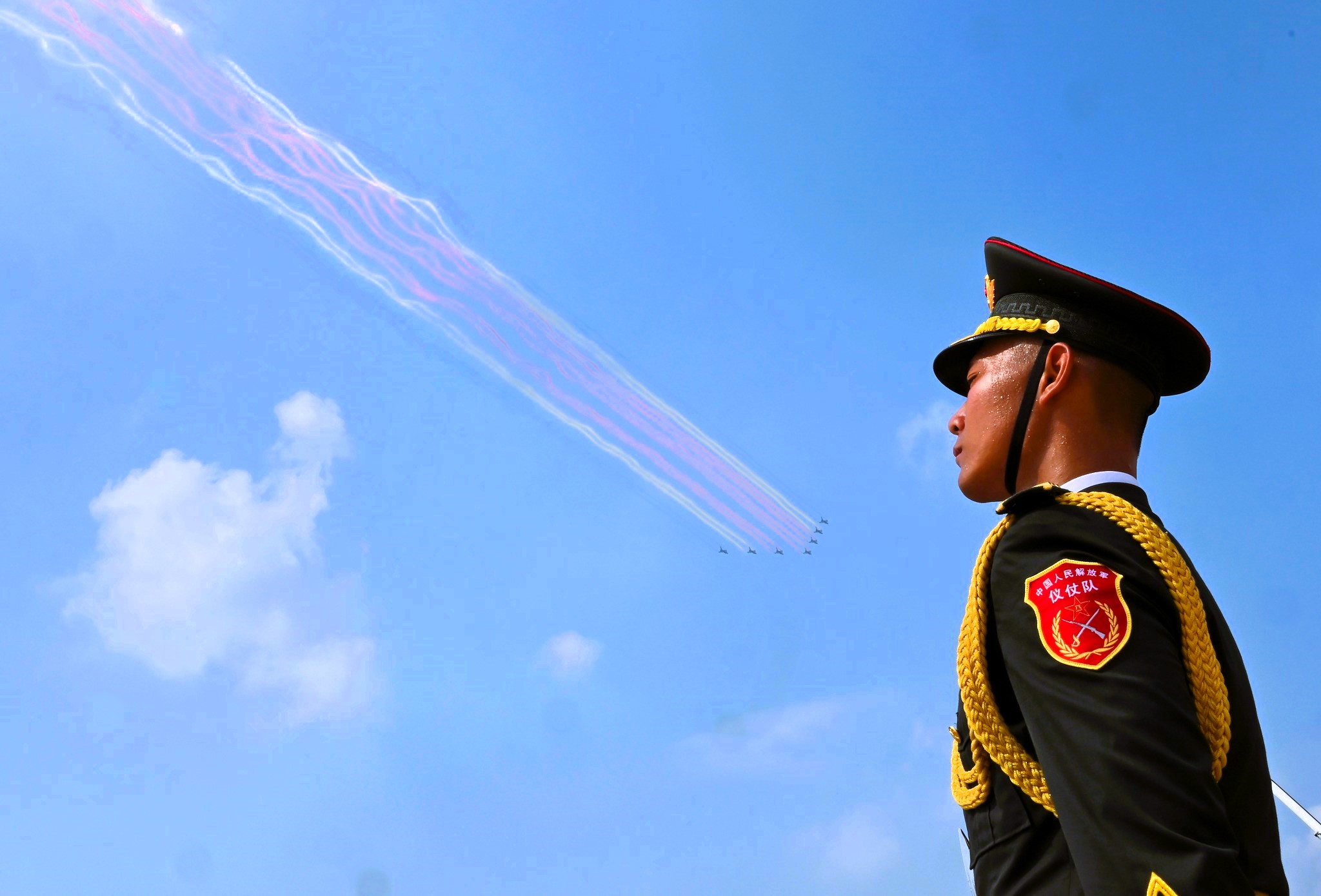 PLA honor guard soldier watches aircraft fly in formation with colored contrails, representing China’s advancing military capabilities