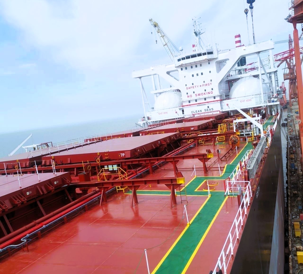 Red and green deck of a bulk carrier vessel docked at port, with another ship visible in the background under overcast skies