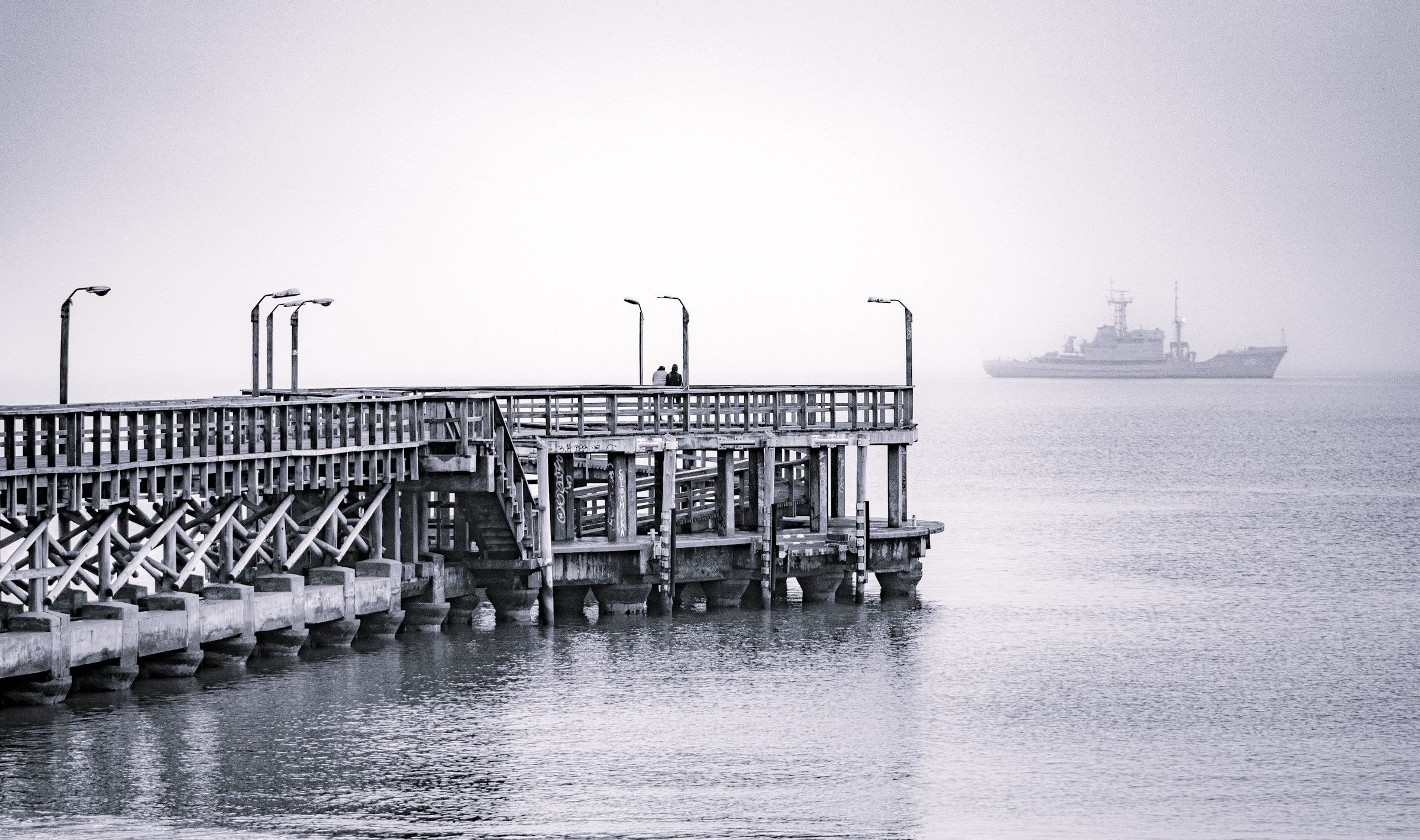 A wooden pier extending over calm water with two people standing at the end and a distant ship in the background