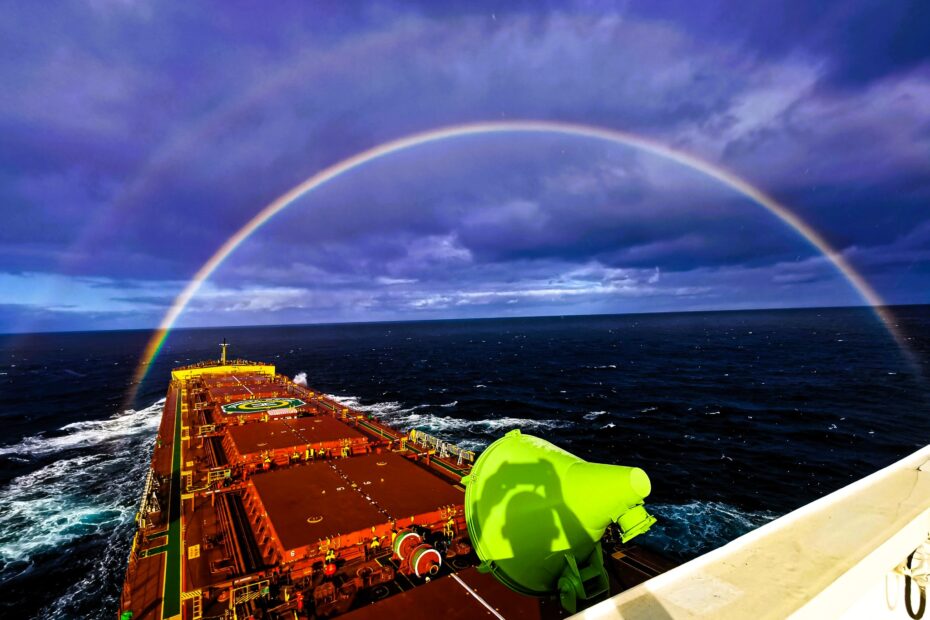 Bulk carrier at sea under a dramatic sky with a rainbow arching over the vessel’s bow
