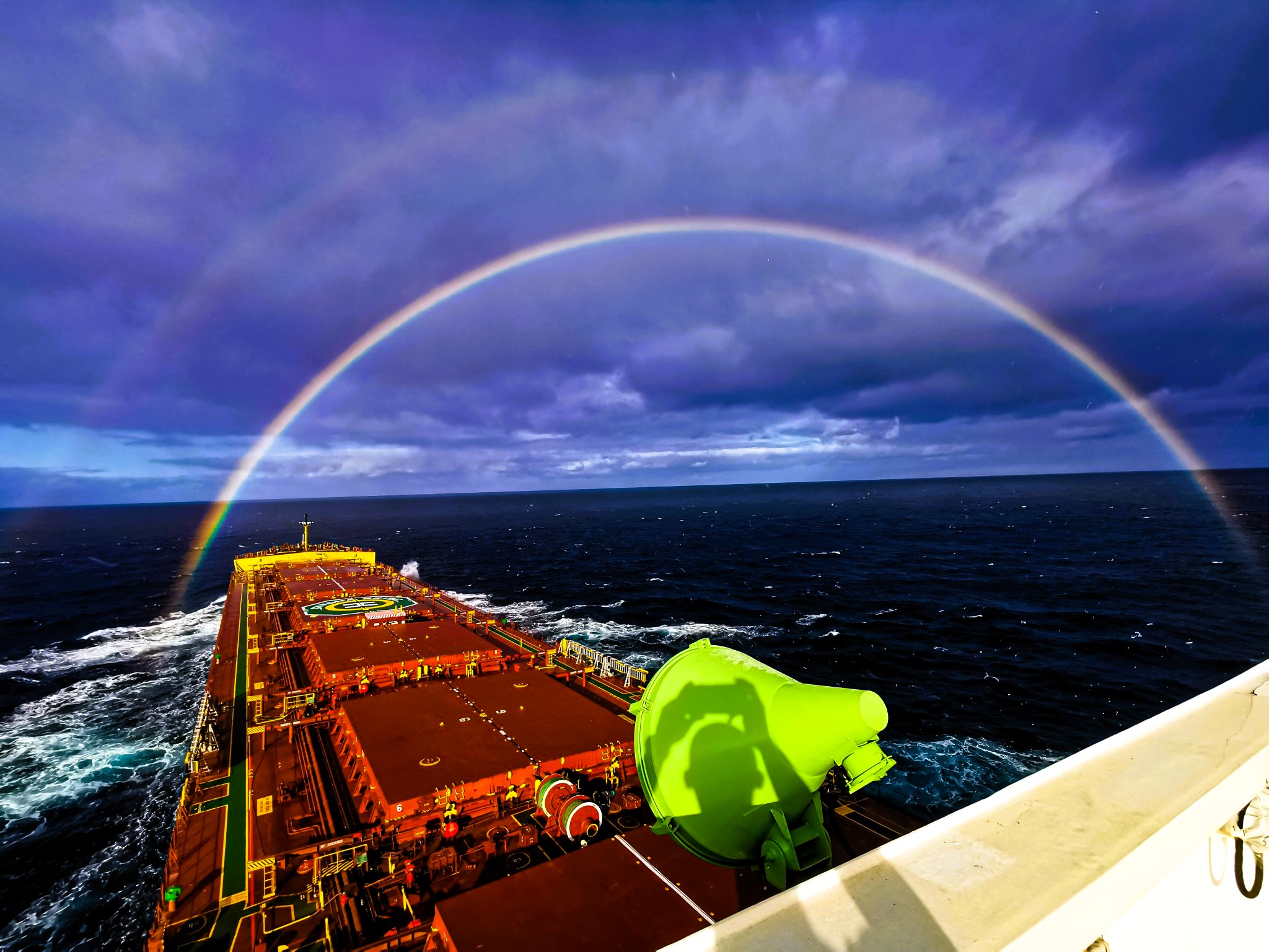 Bulk carrier at sea under a dramatic sky with a rainbow arching over the vessel’s bow