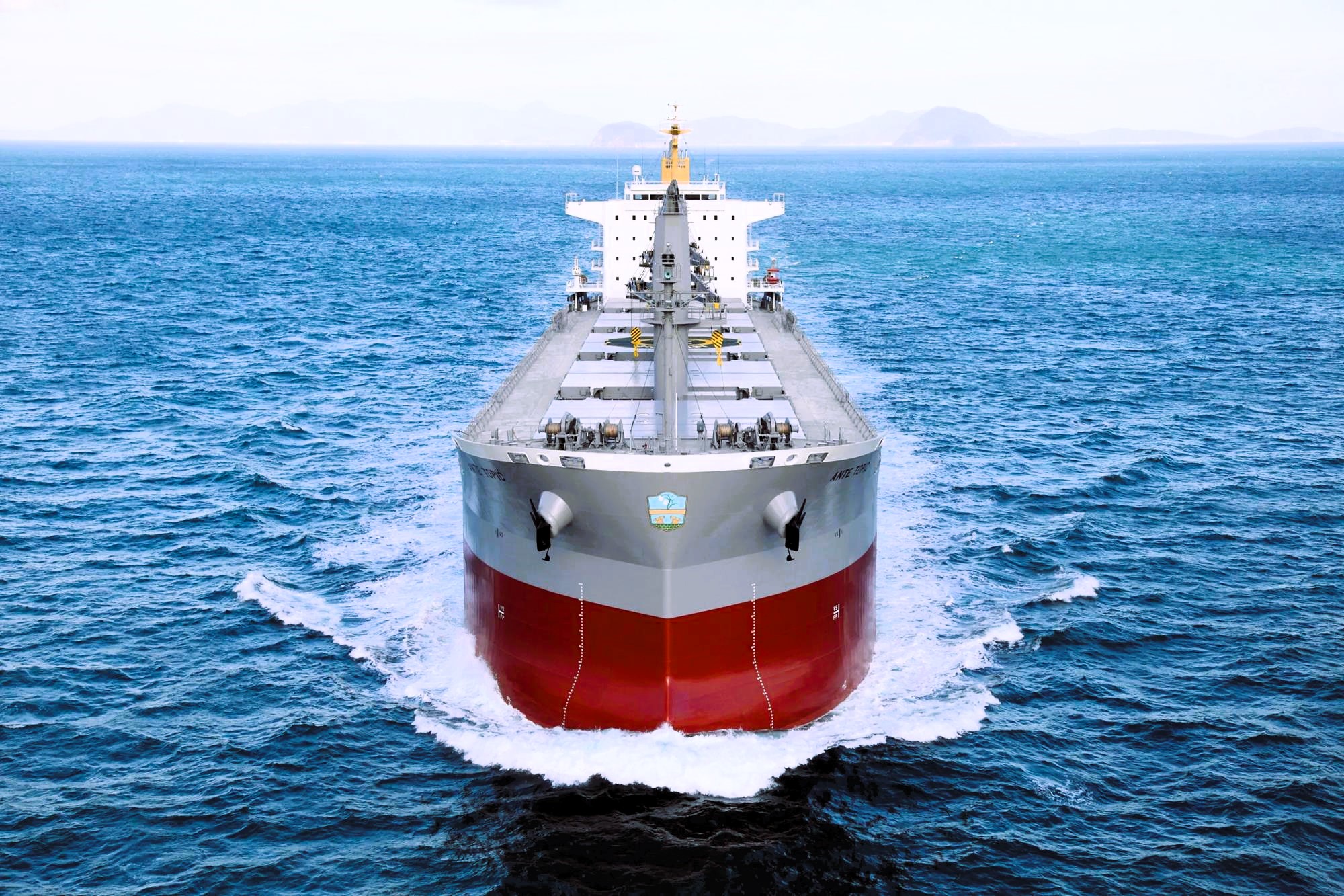 Front-facing bulk carrier sailing through open sea, bow cutting water with horizon visible under a clear sky