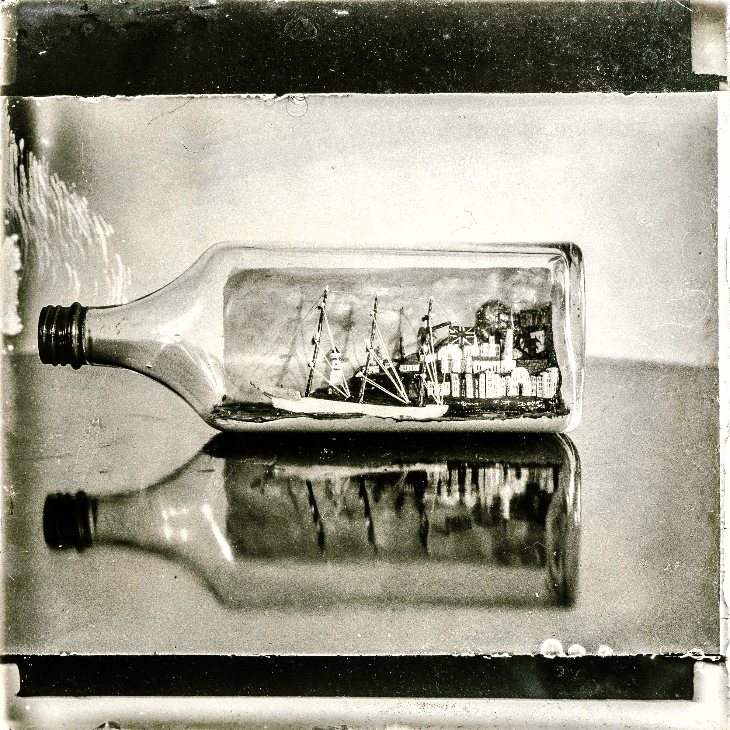 A miniature sailing ship inside a glass bottle, reflected on a dark surface, photographed in vintage black-and-white style