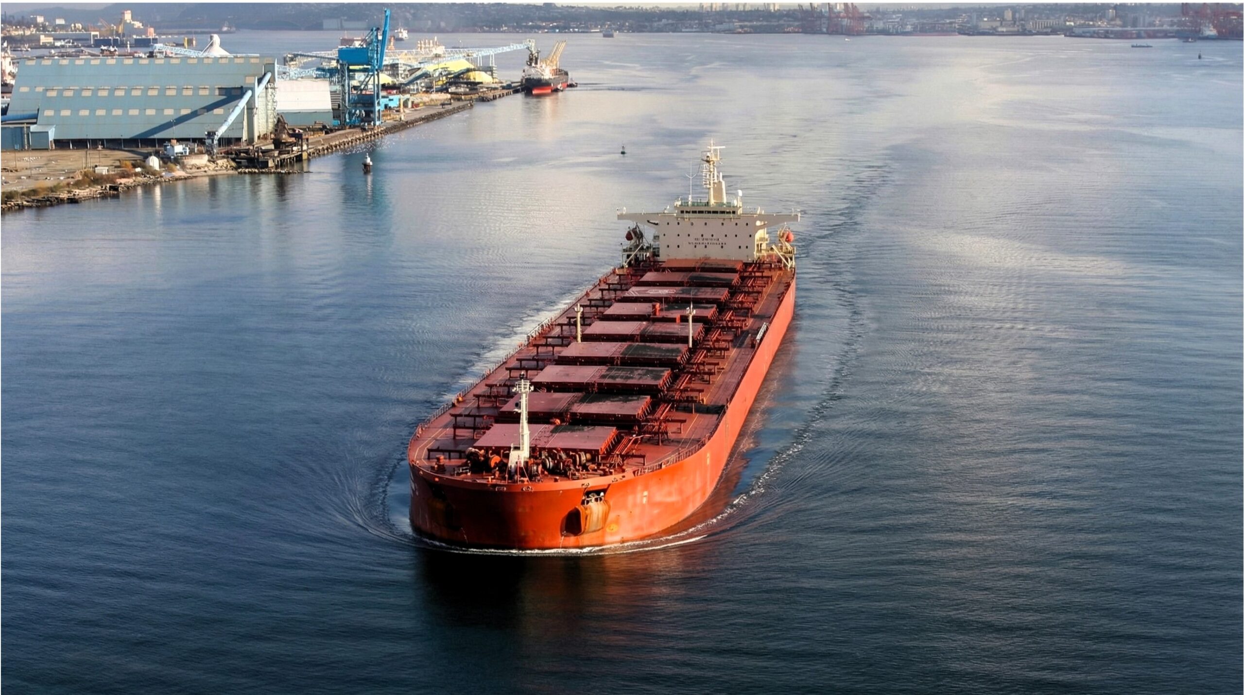Large red bulk carrier sailing in a wide river channel near industrial port facilities and grain terminals under clear daylight skies