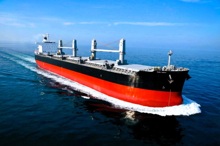 A large bulk carrier vessel sailing across calm open sea, viewed from above, with a red hull and deck cranes visible
