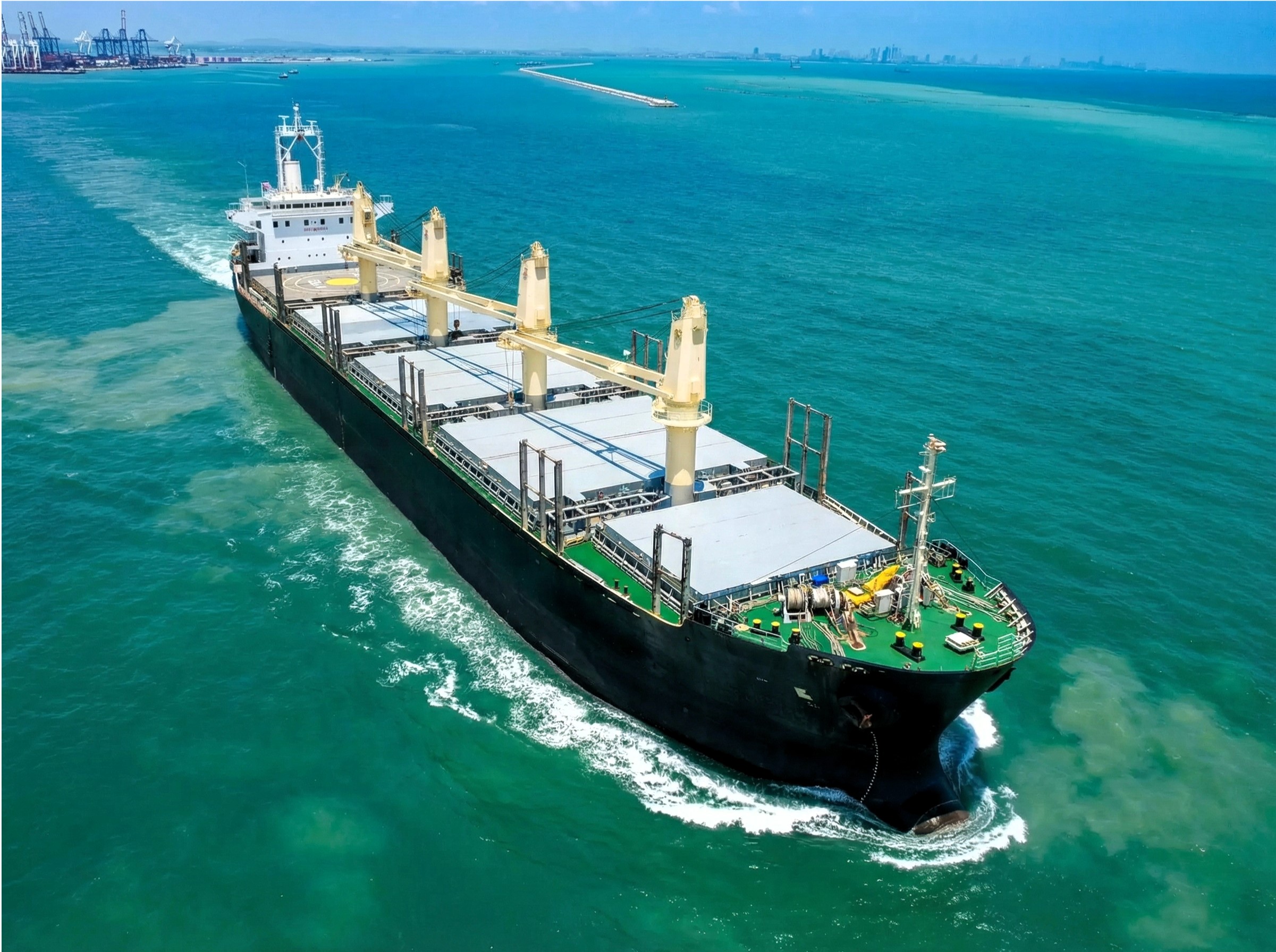 Bulk carrier vessel sailing through turquoise coastal waters near a port area, with cranes and distant city skyline visible