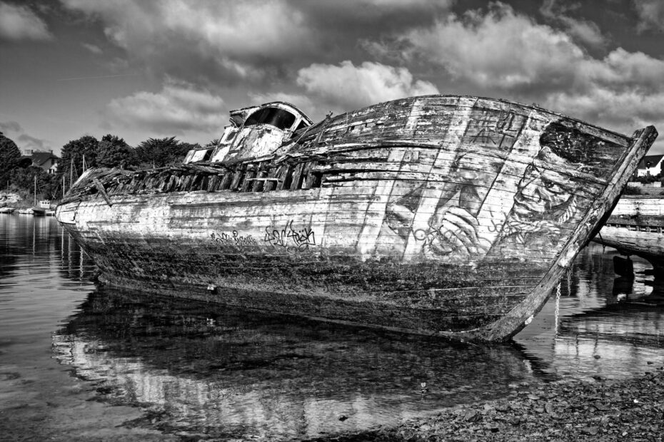 Black-and-white photograph of a decaying wooden boat with graffiti, partially submerged near a quiet shoreline under cloudy skies