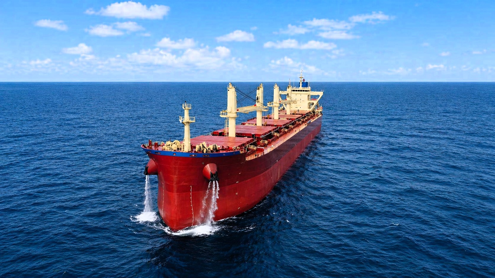 A red bulk carrier sailing in open sea under a partly cloudy sky, viewed from above, with enhanced natural colors and no visible ship name