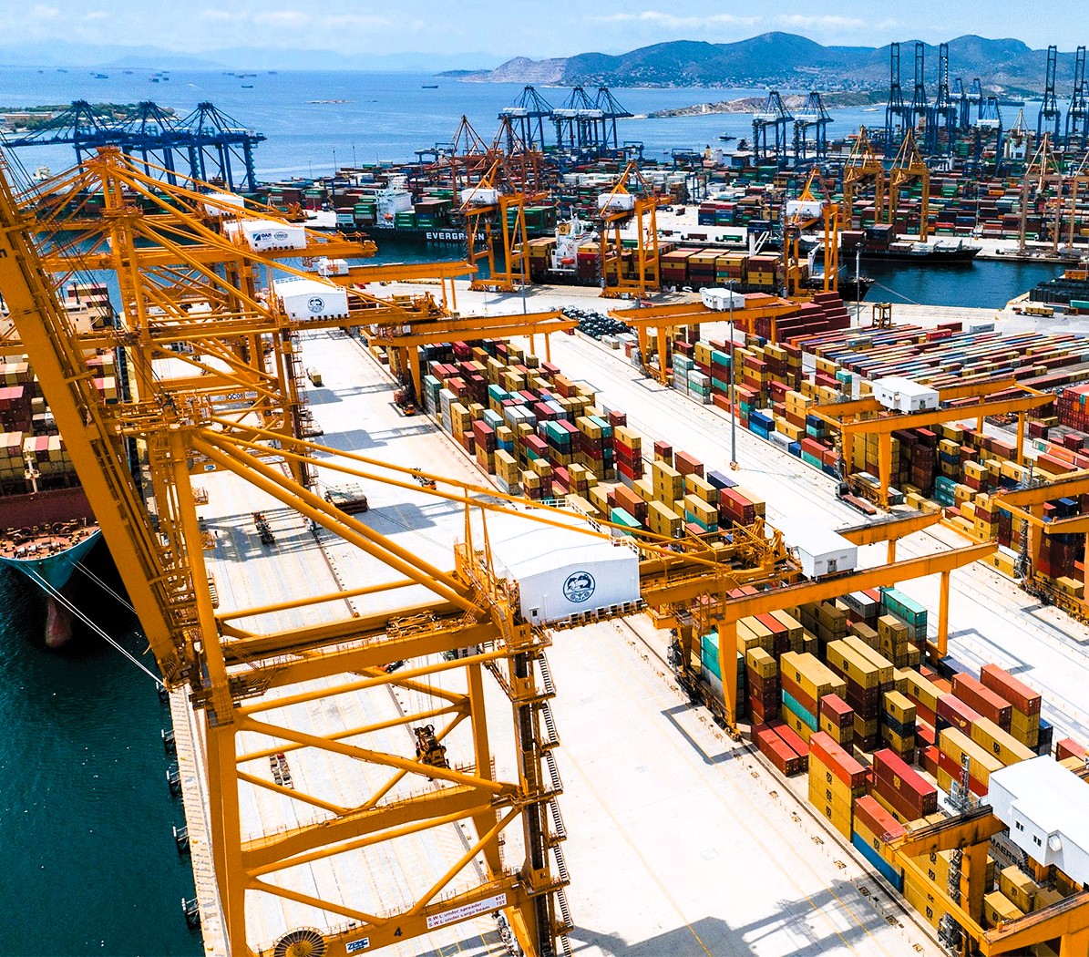 Container terminal with cranes and stacked shipping containers at the Port of Piraeus, overlooking the sea and surrounding hills