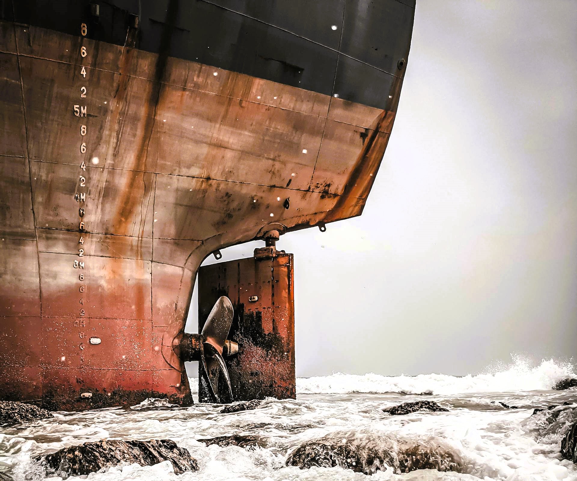 Close-up of a large vessel’s stern, rudder and propeller exposed above churning surf against an overcast sky