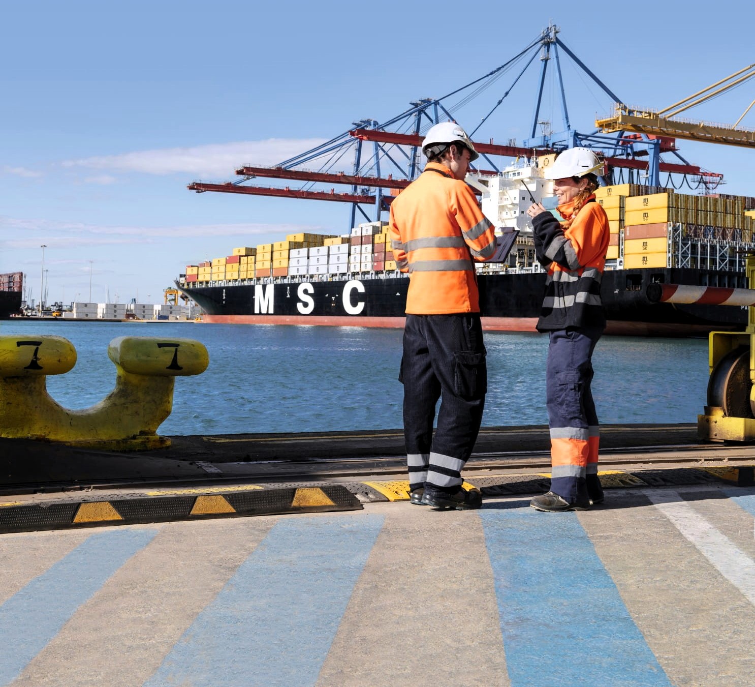 Two port workers in high-visibility gear consult on a quayside as a large MSC container vessel is loaded at berth behind them