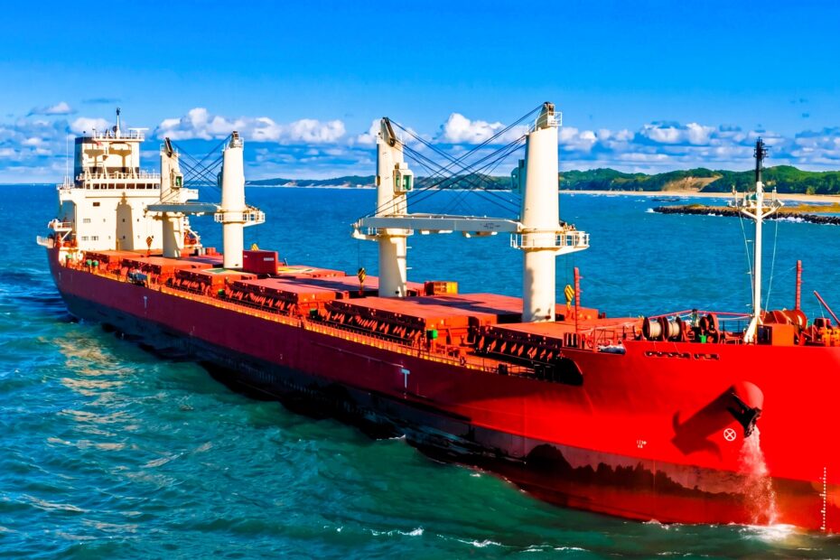 Red bulk carrier sailing in calm blue waters near a coastline under clear sky