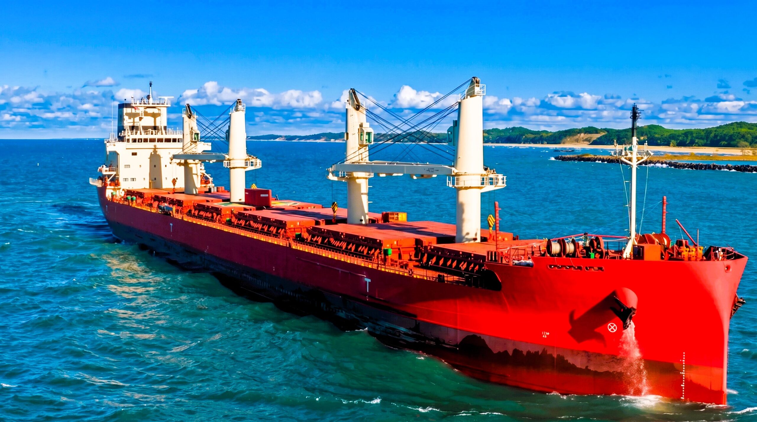Red bulk carrier sailing in calm blue waters near a coastline under clear sky