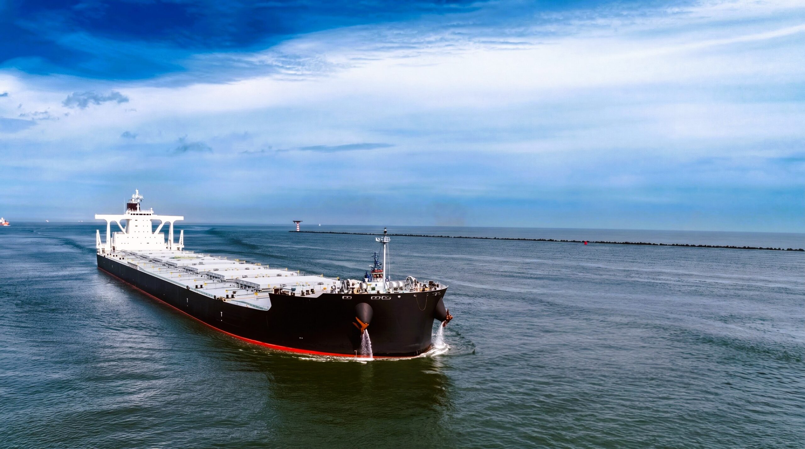 Large Capesize bulk carrier sailing in calm open waters under a partly cloudy sky, approaching a breakwater with a navigation beacon visible in the distance