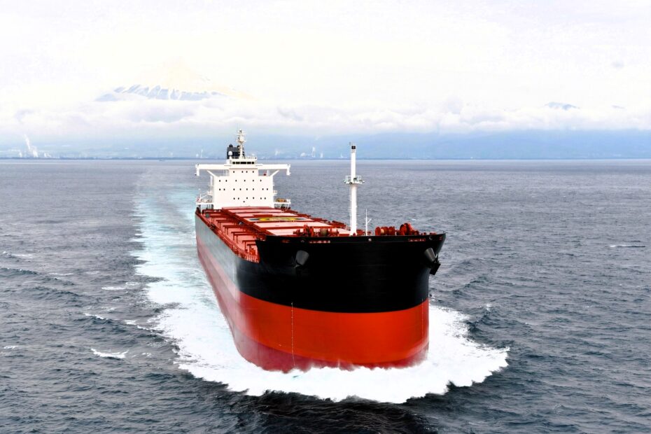 Large bulk carrier sailing through open sea with visible wake, distant mountains and cloudy sky in the background