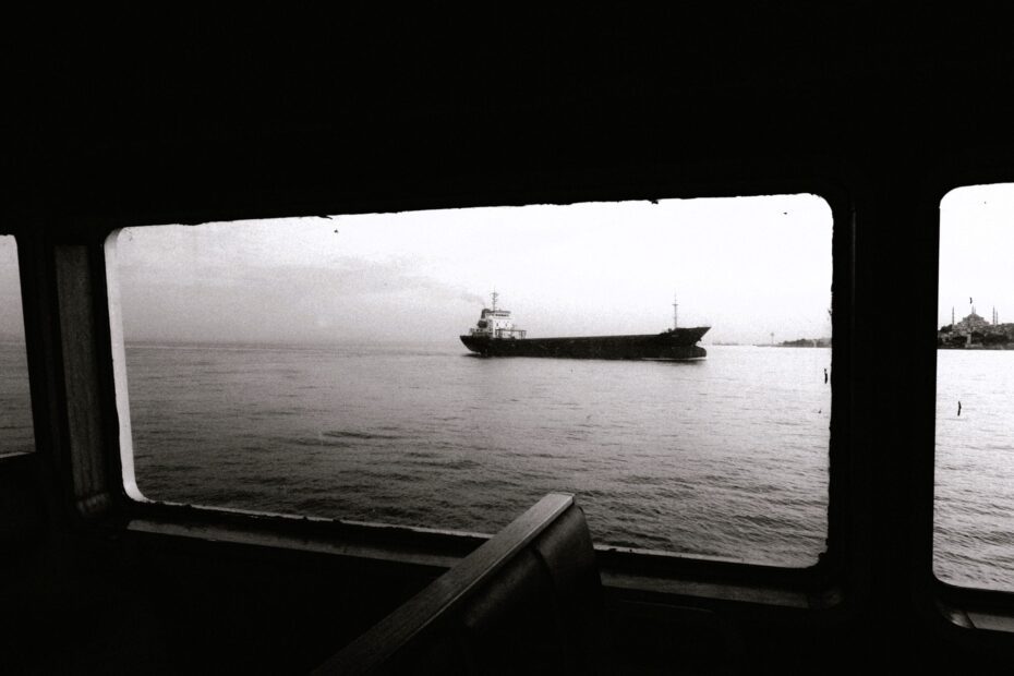 Black-and-white photograph taken from inside a ferry or vessel cabin, showing a cargo ship crossing calm waters near a distant shoreline