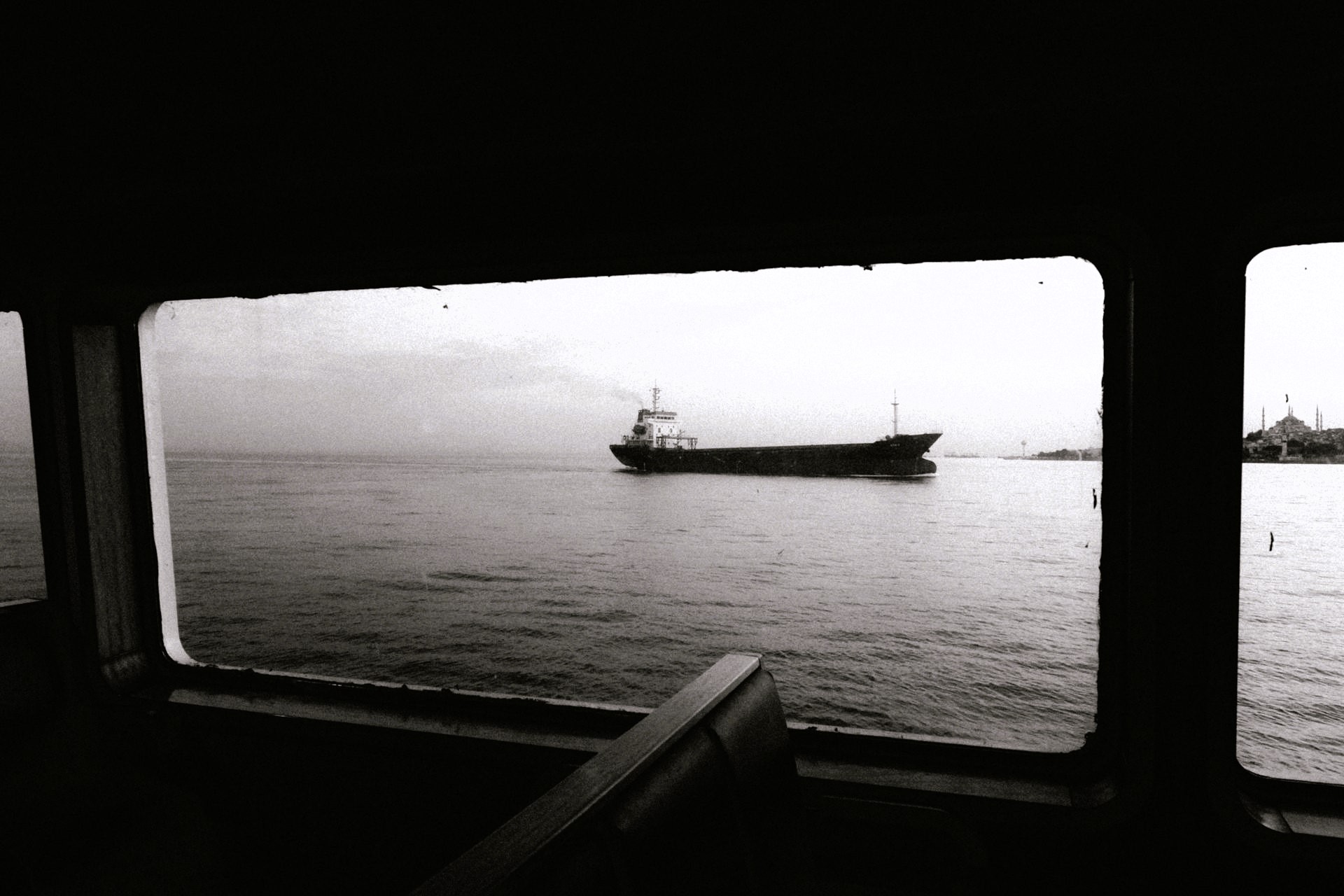 Black-and-white photograph taken from inside a ferry or vessel cabin, showing a cargo ship crossing calm waters near a distant shoreline