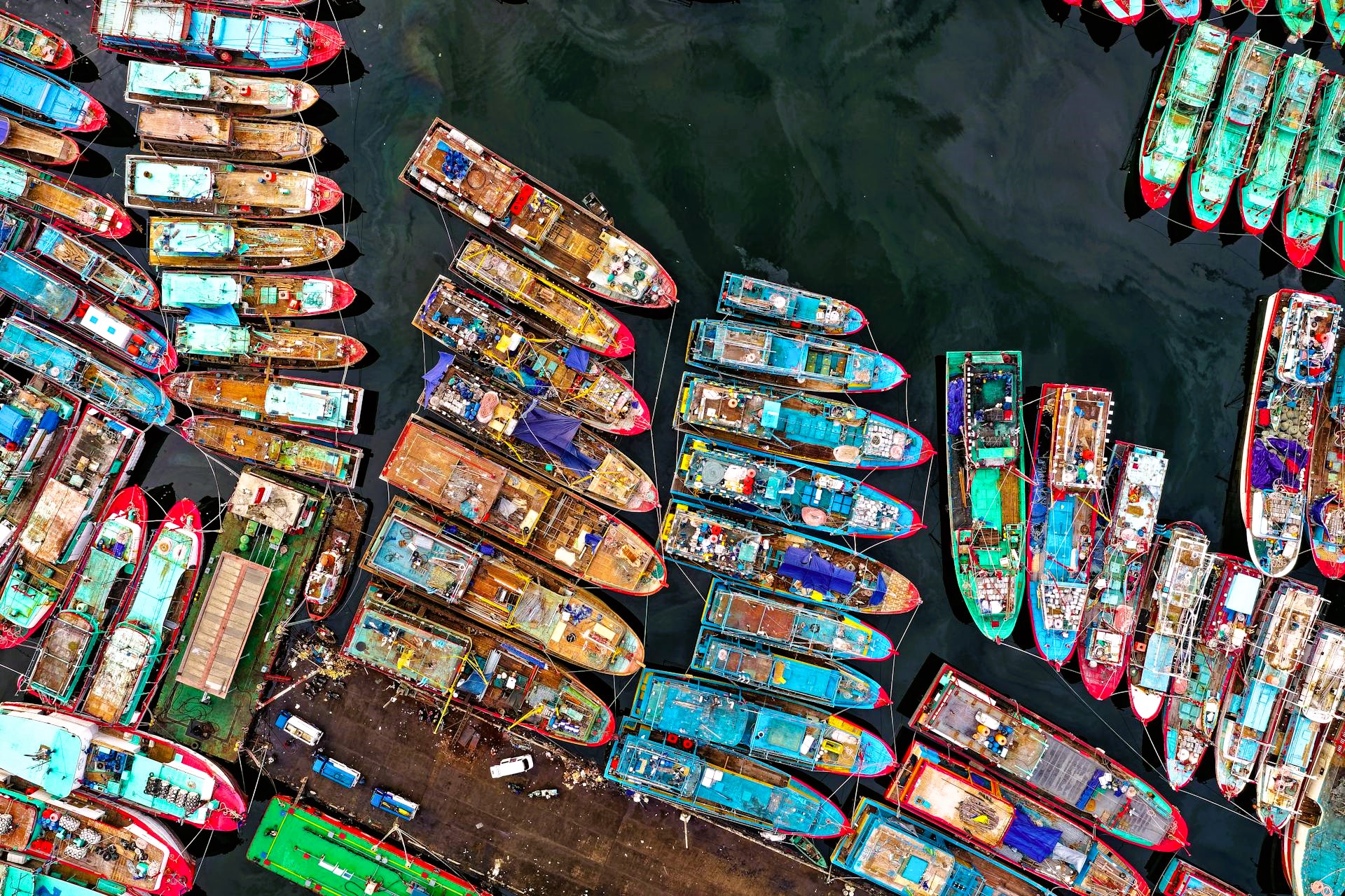 Aerial view of tightly packed colorful vessels moored in dark harbour waters, forming dense geometric patterns across a busy maritime dock