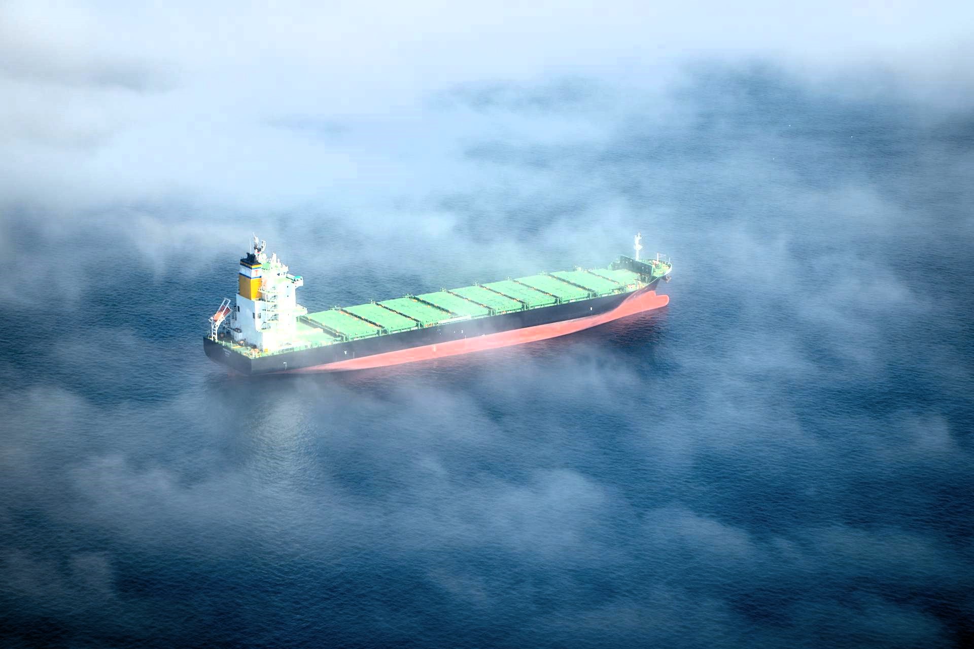 A dry bulk carrier sails through mist-covered open waters, partially obscured by low clouds above a deep blue sea